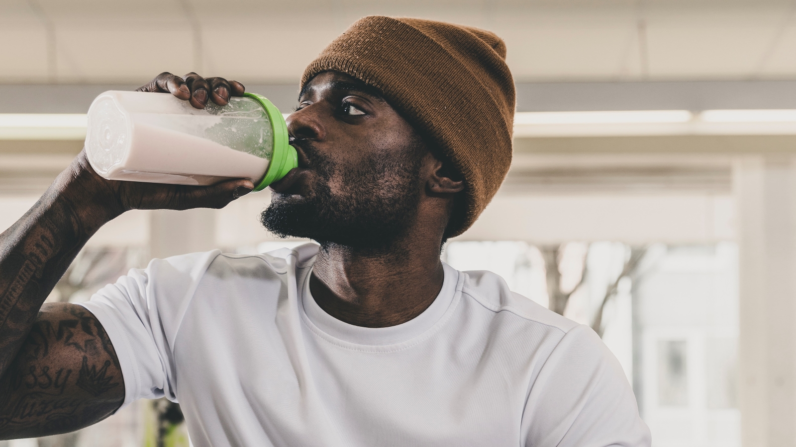 Веган против сывороточного протеина: на фото привлекательный молодой человек в шапке-бини пьет протеиновый коктейль. 