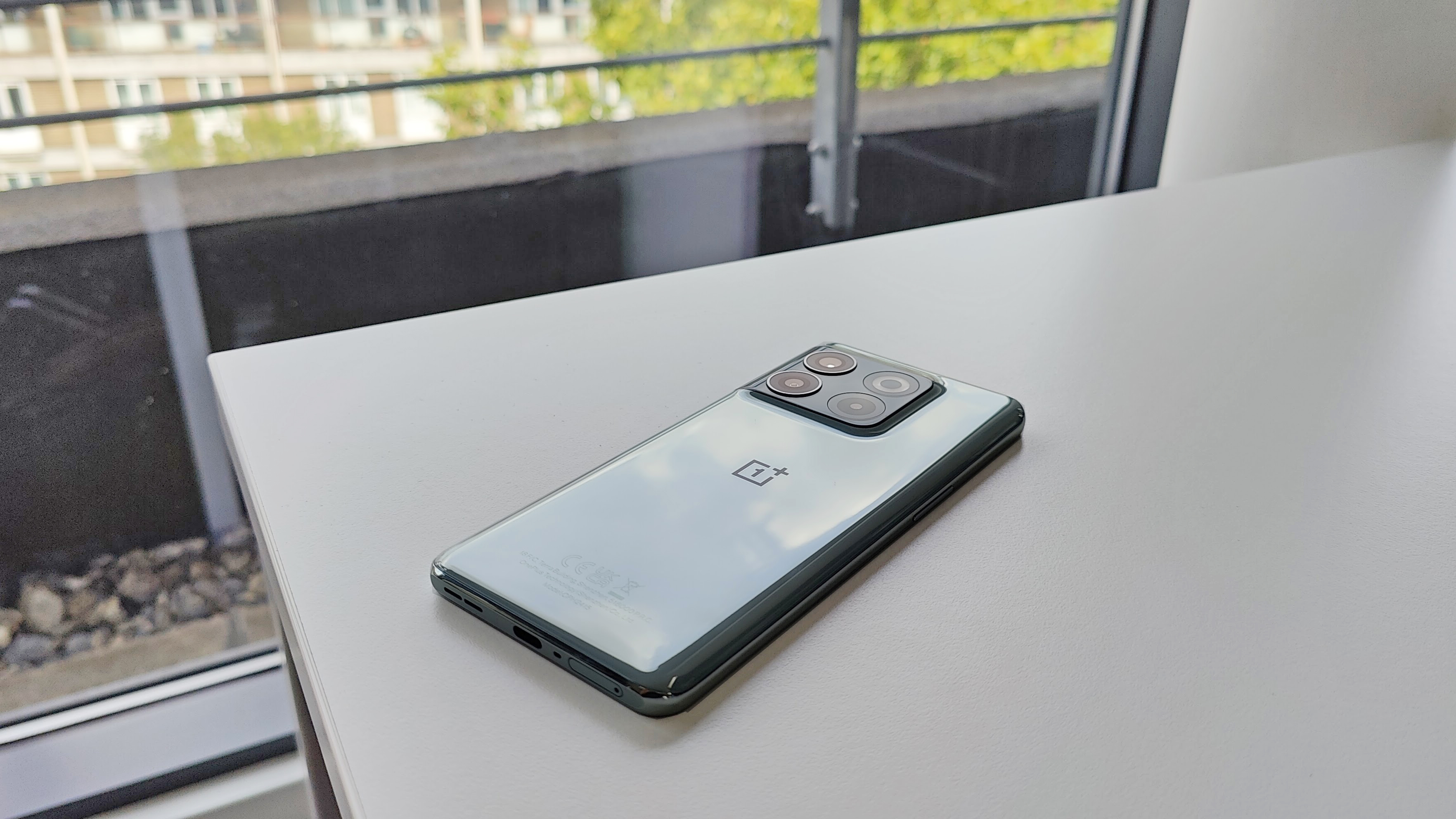 Телефон OnePlus 10T лежит на белом столе с деревьями и зданием позади